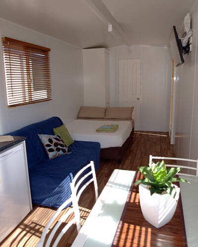 Modern style relocatable cabin for rent with furniture. Featuring a couch, double bed and kitchen table that sits four people. Suitable for a couple looking for cheap accommodation.